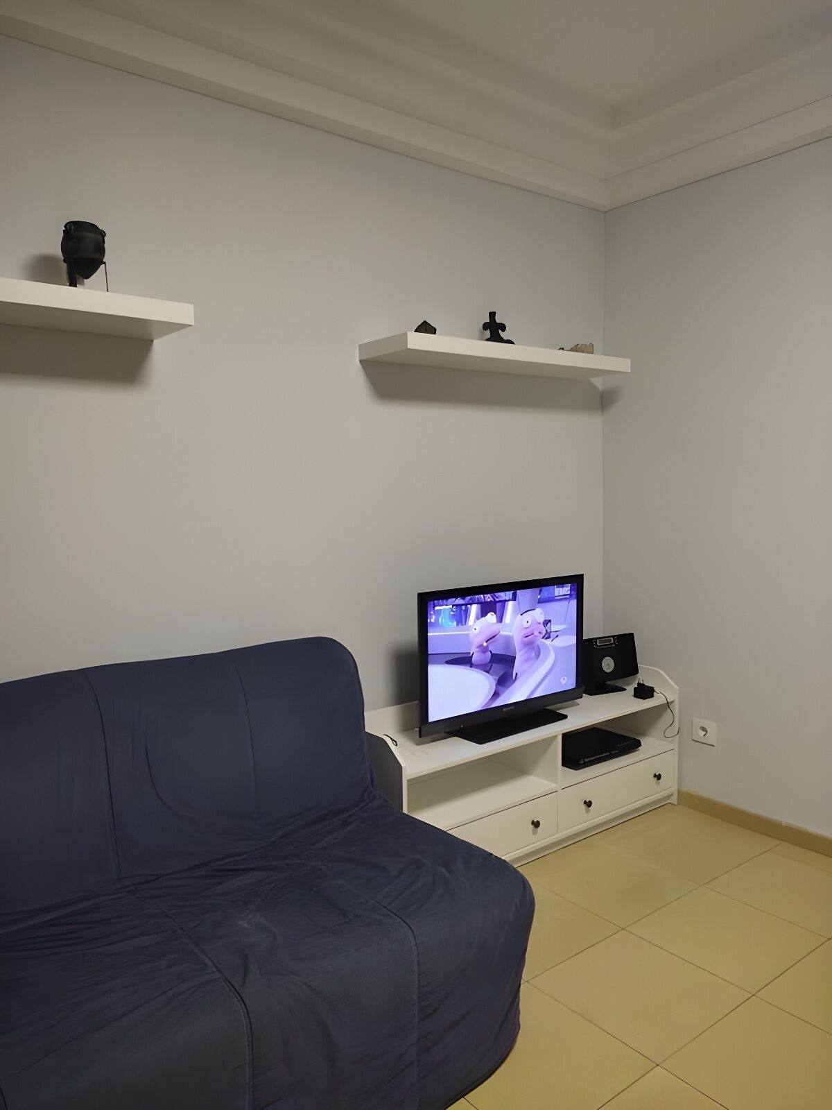 Ett vardagsrum med en mörkblå soffa, en tv på ett vitt tv-bord och två vita hyllor med dekorativa föremål.