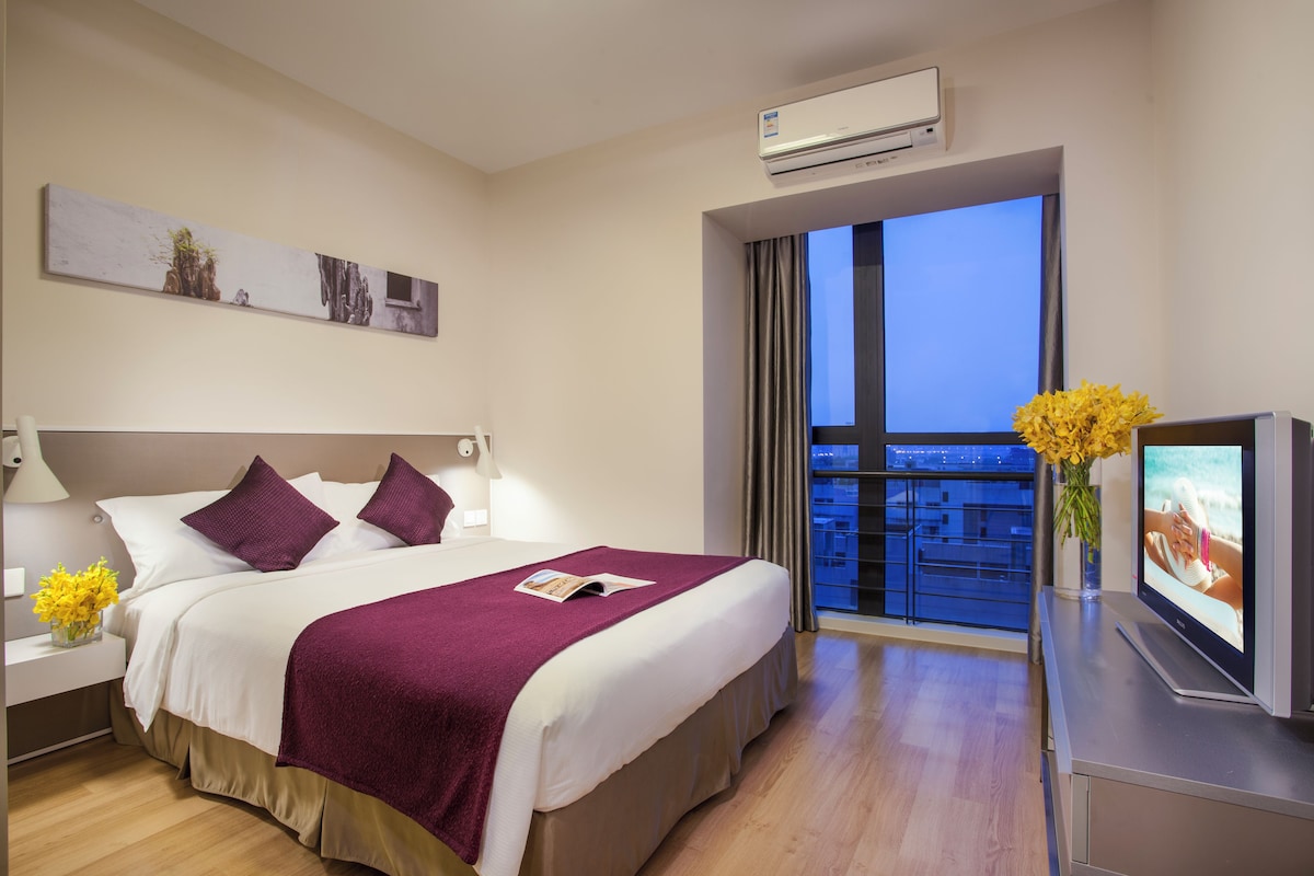 A modern hotel room with a large bed, a flat-screen TV, a window with a city view, and an air conditioning unit.