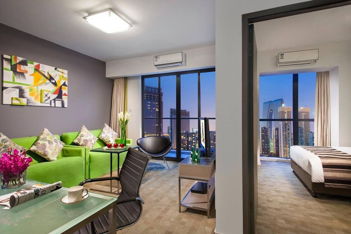 Ein modernes Hotelzimmer mit einem grünen Sofa, einem schwarzen Sessel, einem kleinen Tisch mit Tasse und Zeitschrift sowie einem großen Fenster mit Blick auf die Stadt.