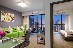 A modern hotel room with a green sofa, a black chair, a small table with a cup and a magazine, a bed, and a large window offering a city view.