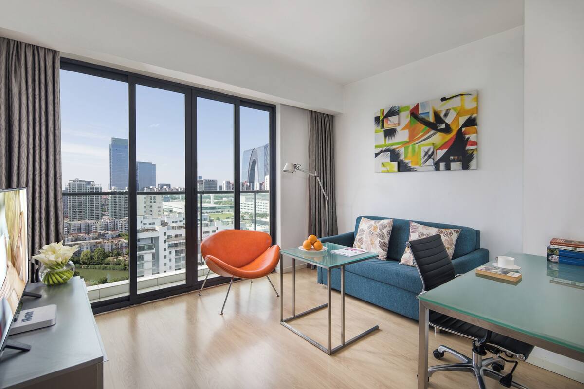 Ein modernes Wohnzimmer mit großem Fenster, einem blauen Sofa, einem orangefarbenen Sessel, einem Glastisch und Blick auf die Stadt.