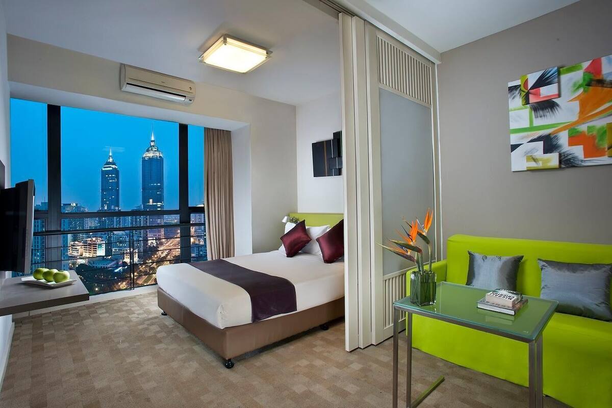 Ein Hotelzimmer mit Bett, Schreibtisch, einem grünen Sofa, einem großen Fenster mit Stadtblick und einem Bild an der Wand.