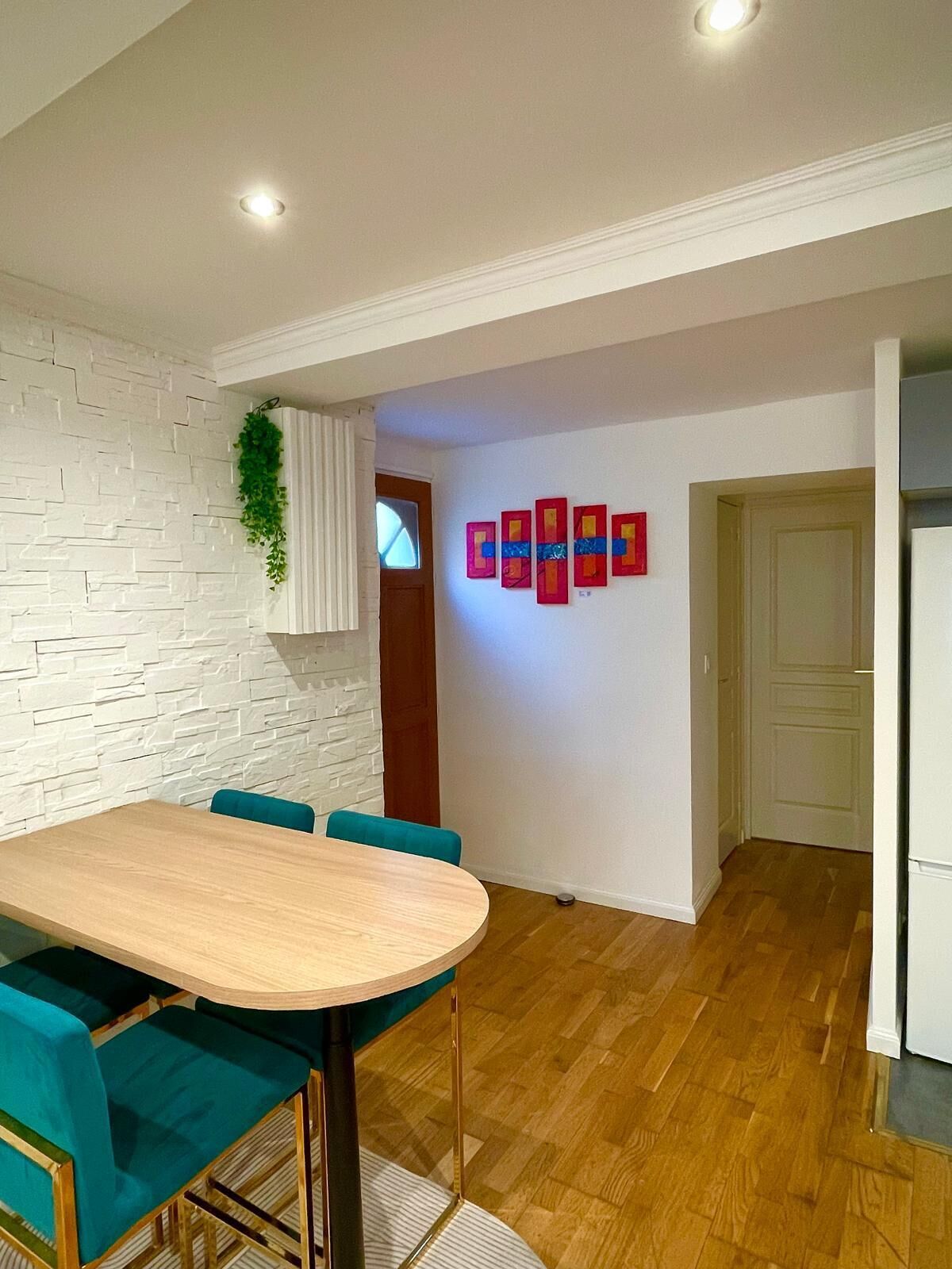 Un coin repas avec une table en bois et des chaises de couleur turquoise, un mur en briques blanches et une œuvre d’art encadrée de rouge accrochée au mur.
