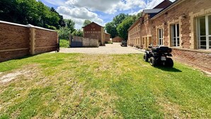 A courtyard with a paved path, brick buildings, and a vehicle. - La Laiterie Gite de groupe Proix (Proix)