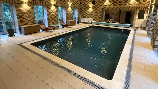 An indoor swimming pool with a tiled floor, a poolside seating area, and a patterned wall. - La Laiterie Gite de groupe Proix (Proix)