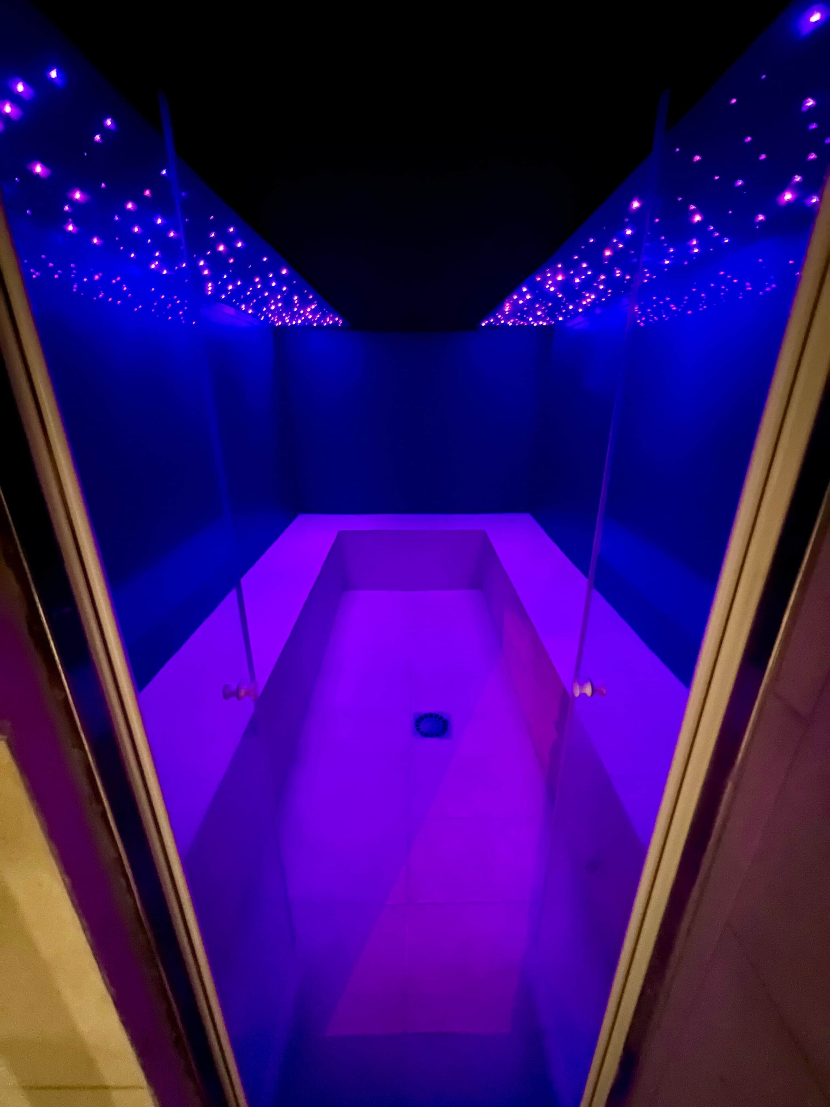A narrow, dimly lit room with a central drain and walls adorned with small, evenly spaced lights.