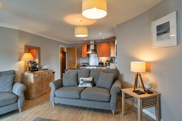 A living room with a grey sofa, a wooden cabinet, and a small table with a lamp. - Apartment in Watergate Bay With Sea Views (Newquay)