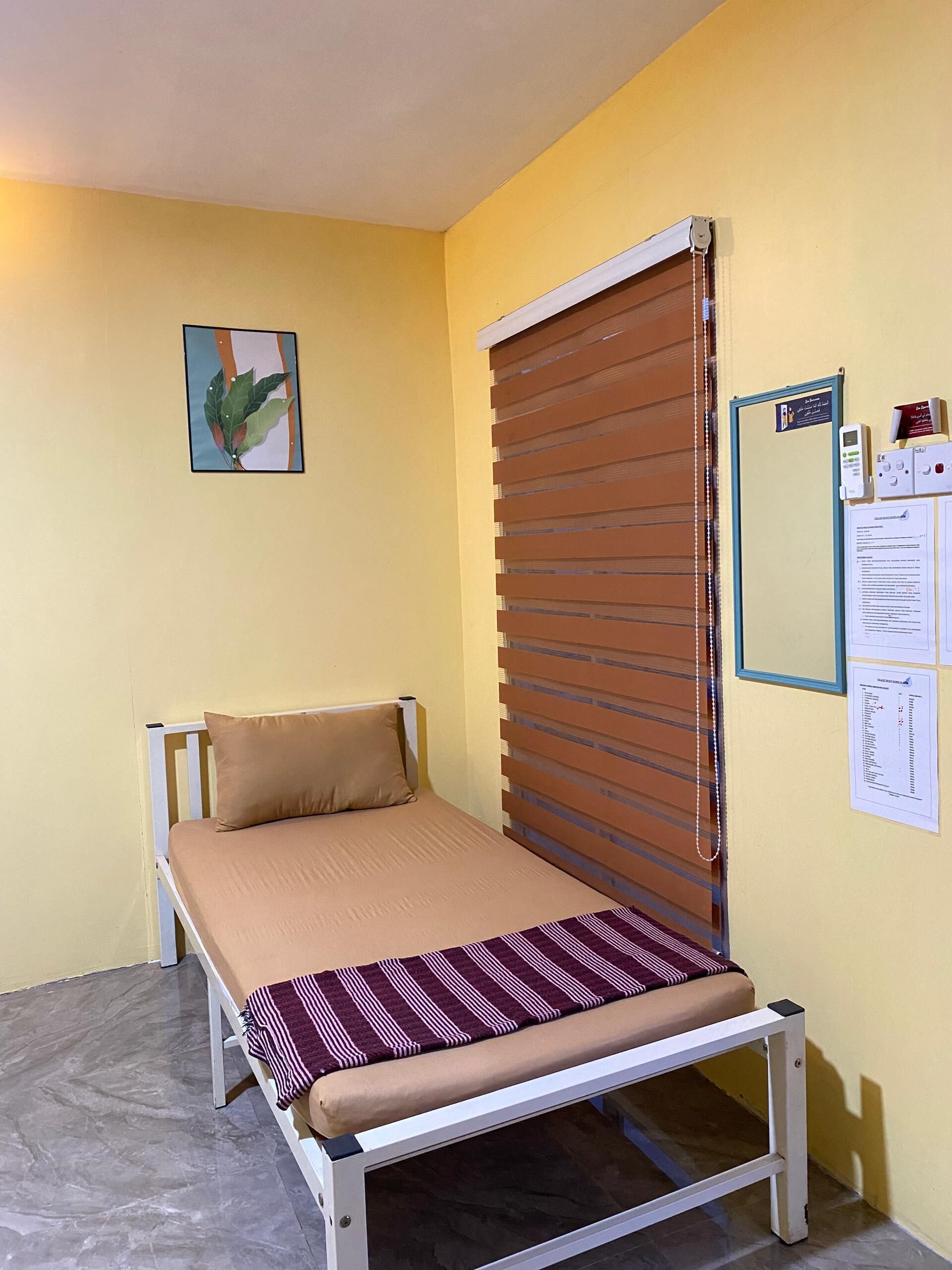 A small room with a single bed, a window with blinds, a wall-mounted thermostat, and a framed picture.