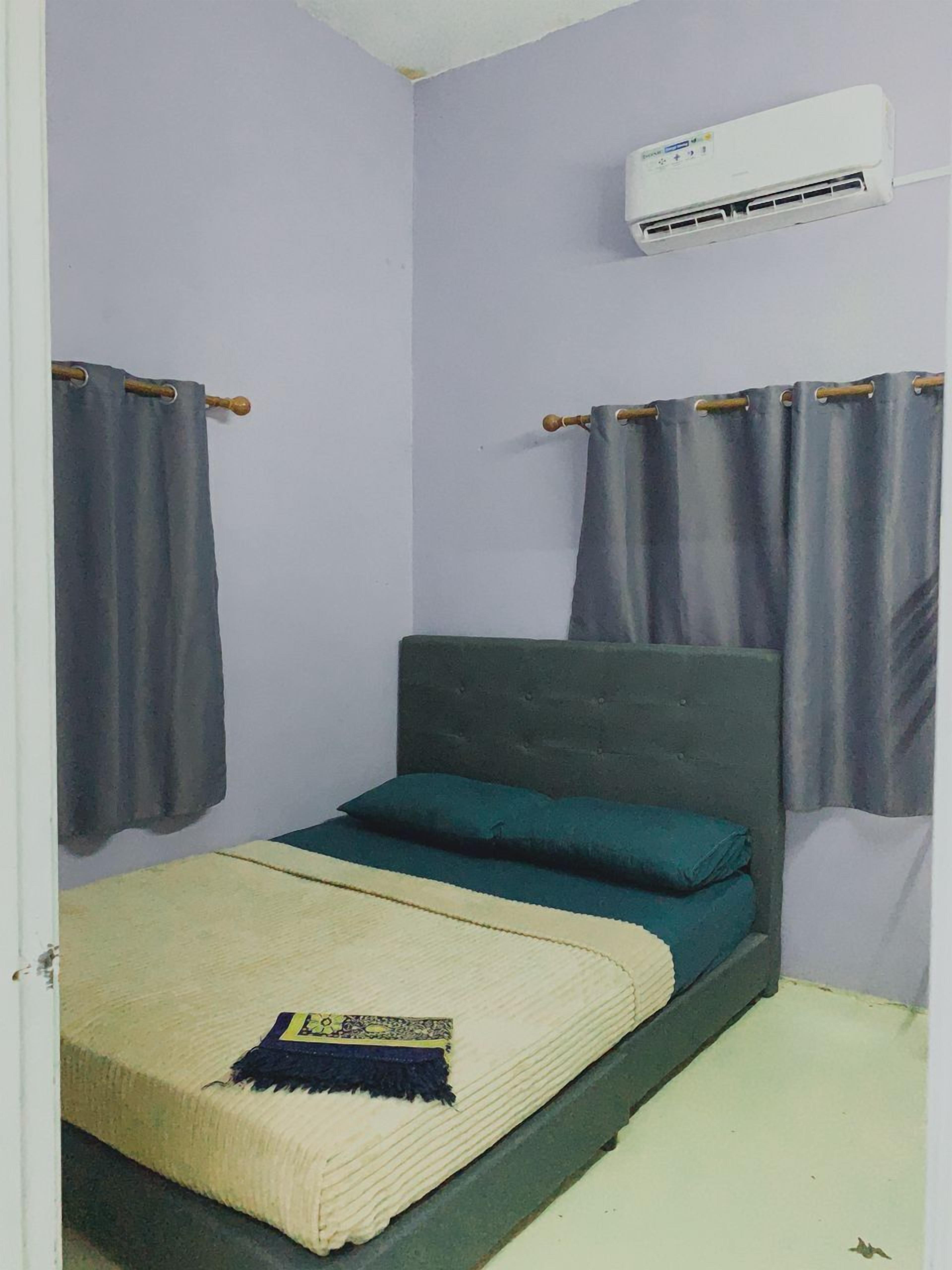 A small, single-bed room with a grey headboard, a bed with a beige blanket and a blue throw, grey curtains, and a white air conditioning unit mounted on the wall.