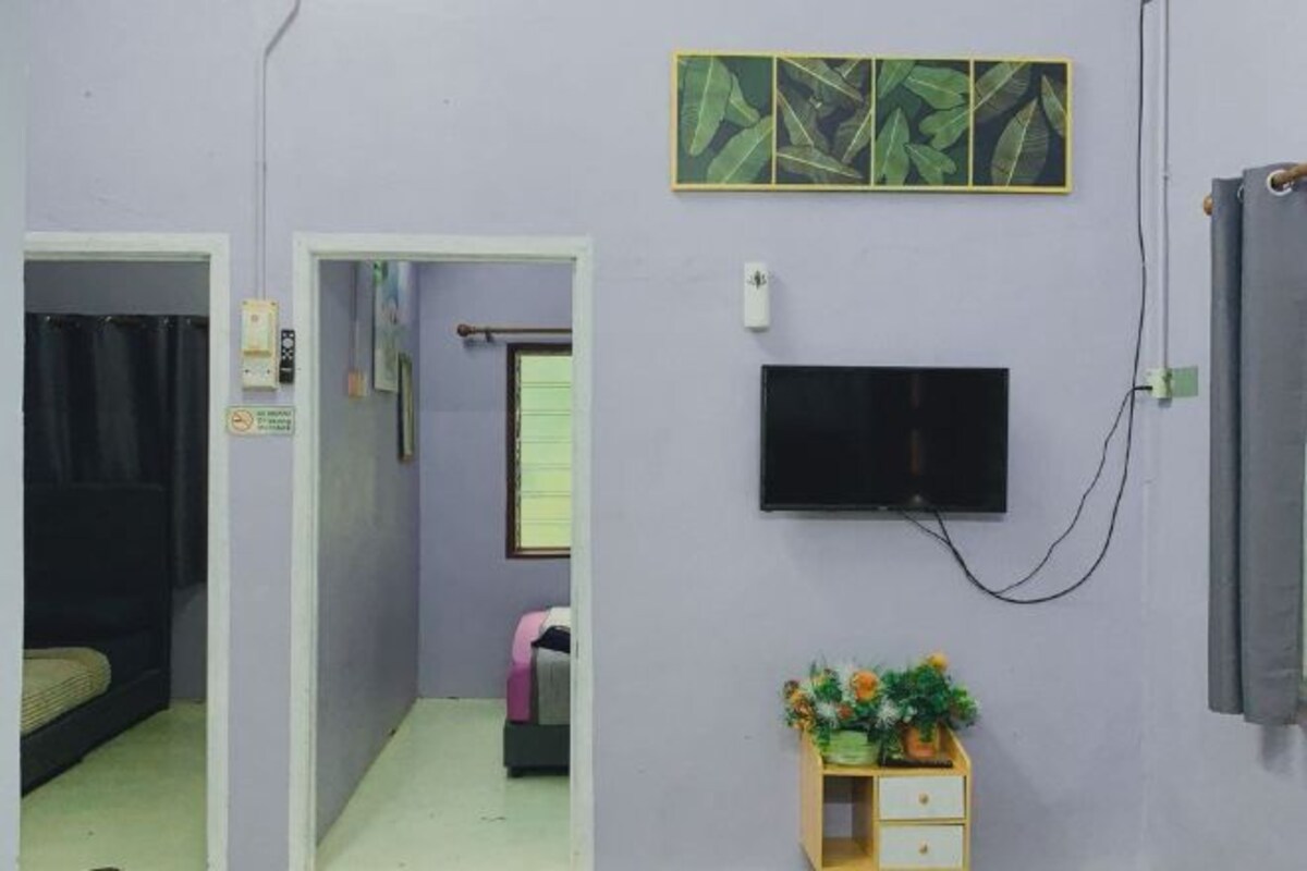 Een kamer met een bed, een aan de muur bevestigde televisie, een klein houten kastje met bloemen en een deur naar een andere kamer.