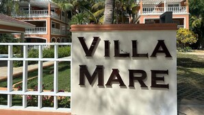 A sign with "VILLA MARE" written on it, surrounded by a white fence, palm trees, and a building with a red roof.