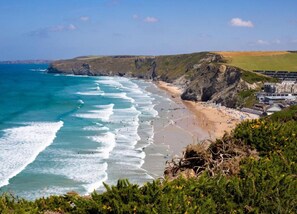 A coastal beach with waves, a sandy shore, and a cliff with vegetation. - Cottage in St Mawgan Near Mawgan Porth Beach (Newquay)