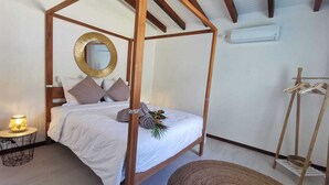 A four-poster bed with a white bedspread and pillows, a round mirror on the wall, a small round table with a lamp, and a wooden chair. - 2 minutes from the beach, pool, jacuzzi and parking included in a secure residence! (Les Trois-Îlets)