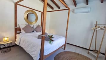A four-poster bed with a white bedspread and pillows, a round mirror on the wall, a small round table with a lamp, and a wooden chair.