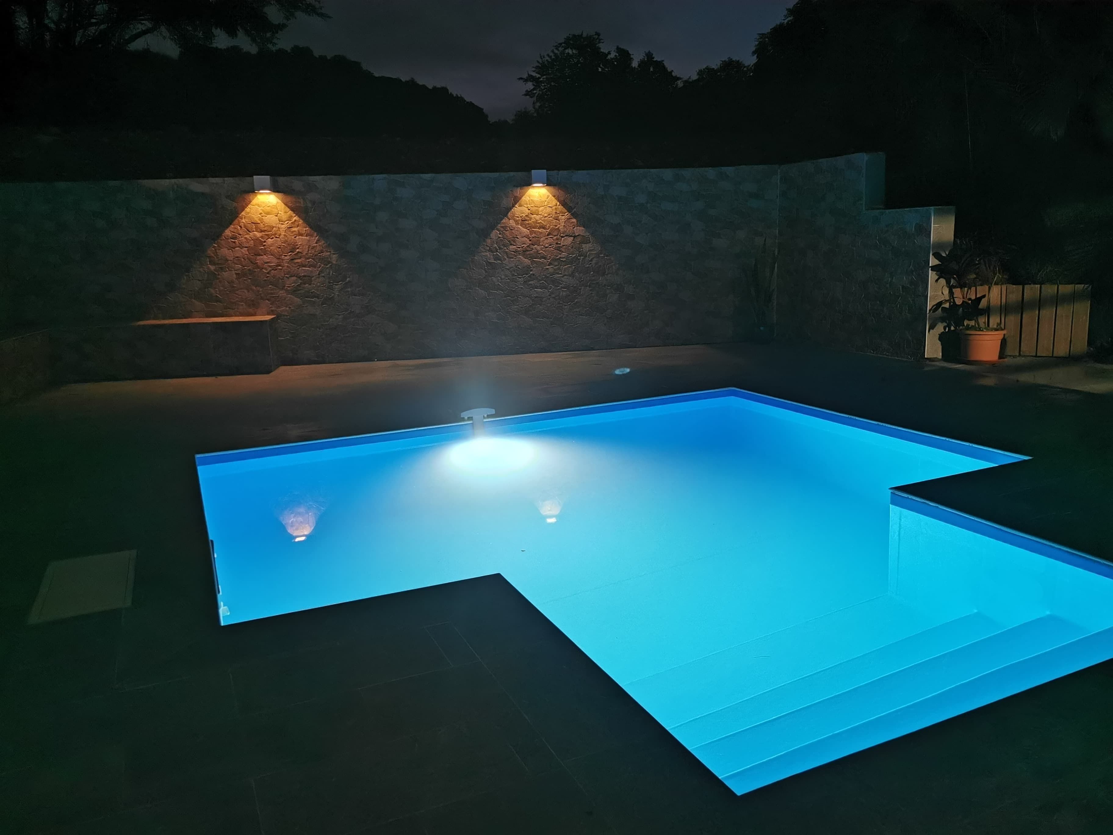 A rectangular swimming pool with a clear blue water surface, surrounded by a stone wall and illuminated by two wall-mounted lights.