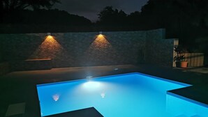A rectangular swimming pool with a clear blue water surface, surrounded by a stone wall and illuminated by two wall-mounted lights. - 2 minutes from the beach, pool, jacuzzi and parking included in a secure residence! (Les Trois-Îlets)