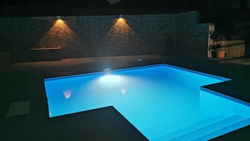 A rectangular swimming pool with a clear blue water surface, surrounded by a stone wall and illuminated by two wall-mounted lights.