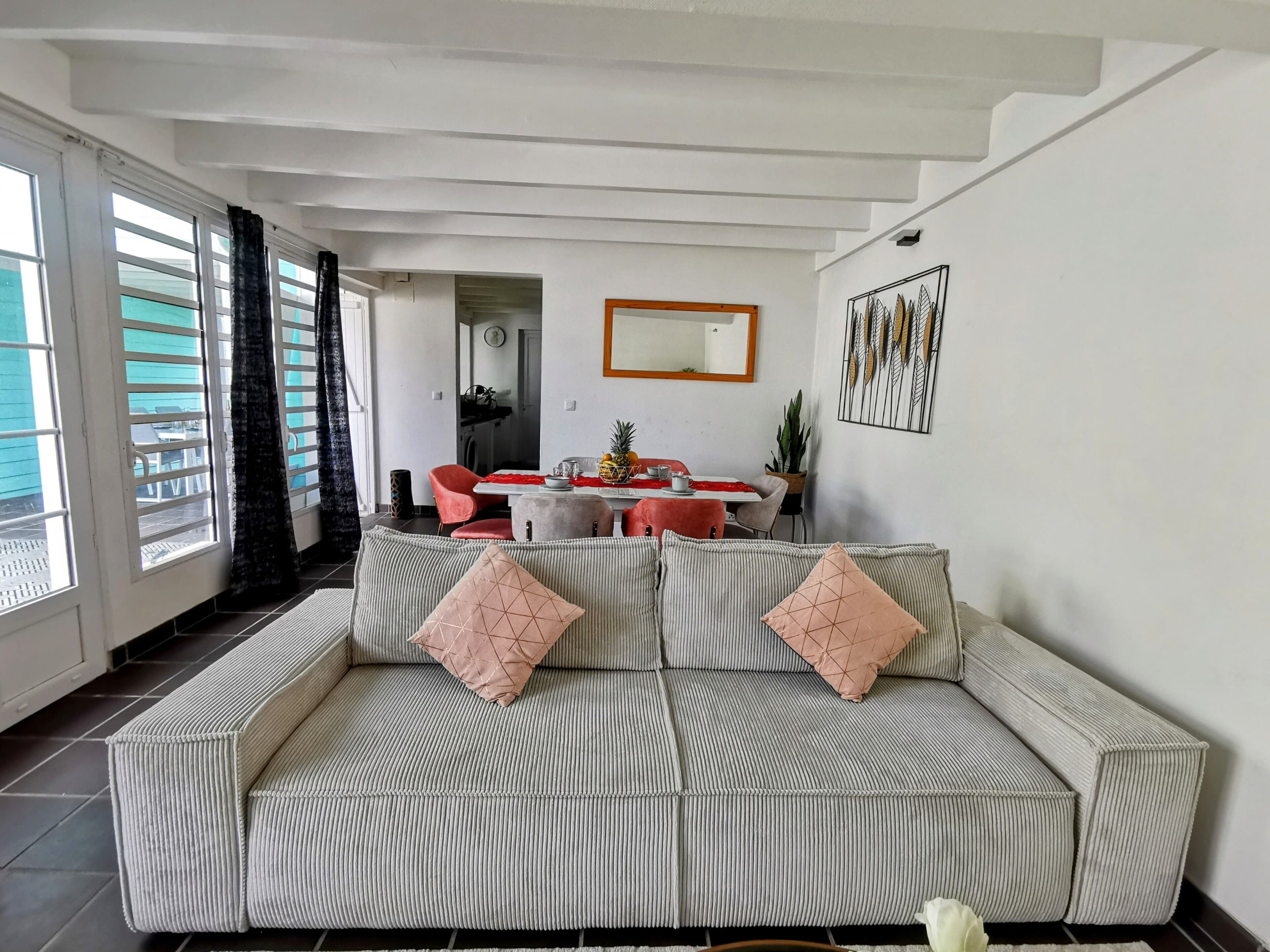 Una sala de estar con un sofá a rayas, una mesa de comedor con sillas, una cocina al fondo y un gran ventanal con cortinas.