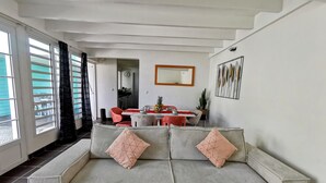 A living room with a striped sofa, a dining table with chairs, a kitchen in the background, and a large window with curtains. - 2 minutes from the beach, pool, jacuzzi and parking included in a secure residence! (Les Trois-Îlets)