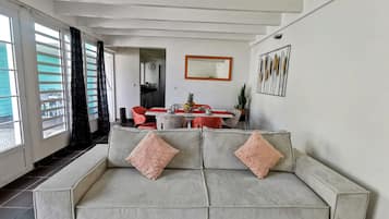 Una sala de estar con un sofá a rayas, una mesa de comedor con sillas, una cocina al fondo y un gran ventanal con cortinas.