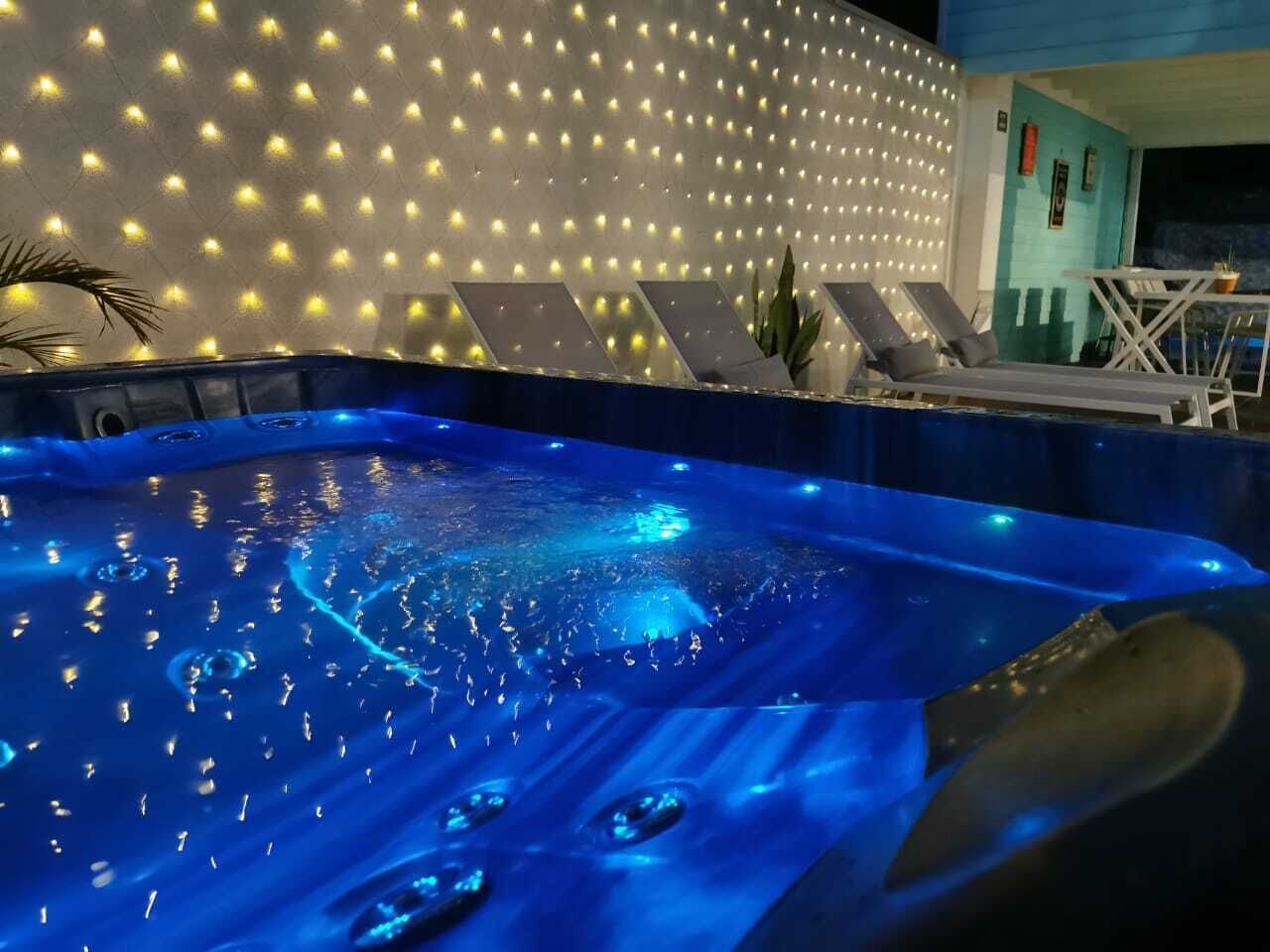 Un jacuzzi con iluminación azul en su interior, rodeado por una pared de luces y una zona de estar con sillones reclinables.