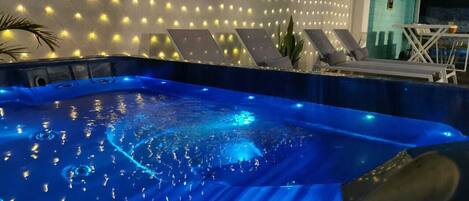 A hot tub with a blue-lit interior, surrounded by a wall of lights and a seating area with lounge chairs.