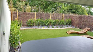 Eine Terrasse mit einem schwarzen Tisch und zwei Holzliegen, umgeben von einem Garten mit Hecke und Steinmauer.