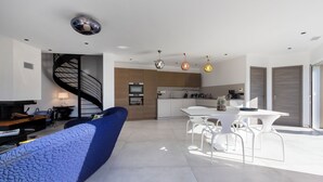 A modern living room with a blue textured sofa, a white dining area with a table and chairs, a kitchen with wooden cabinets, and a spiral staircase. - The beach house (Calcatoggio)