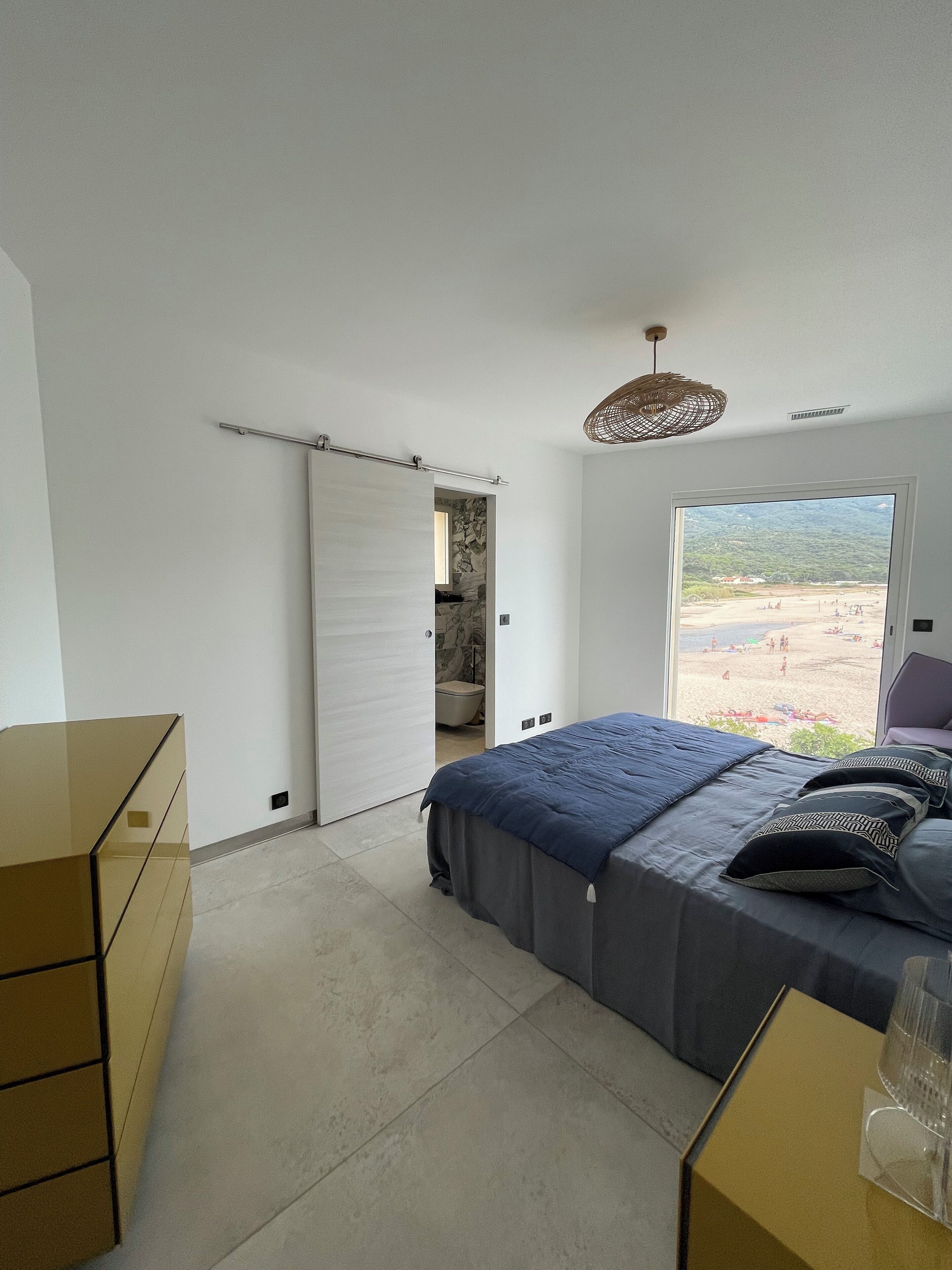 A bedroom with a bed, a dresser, a window with a beach view, and a door leading to another room.
