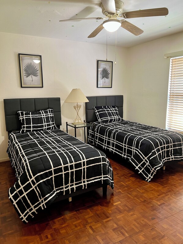 Una habitación con dos camas, ventilador de techo y cuadros con hojas en las paredes.