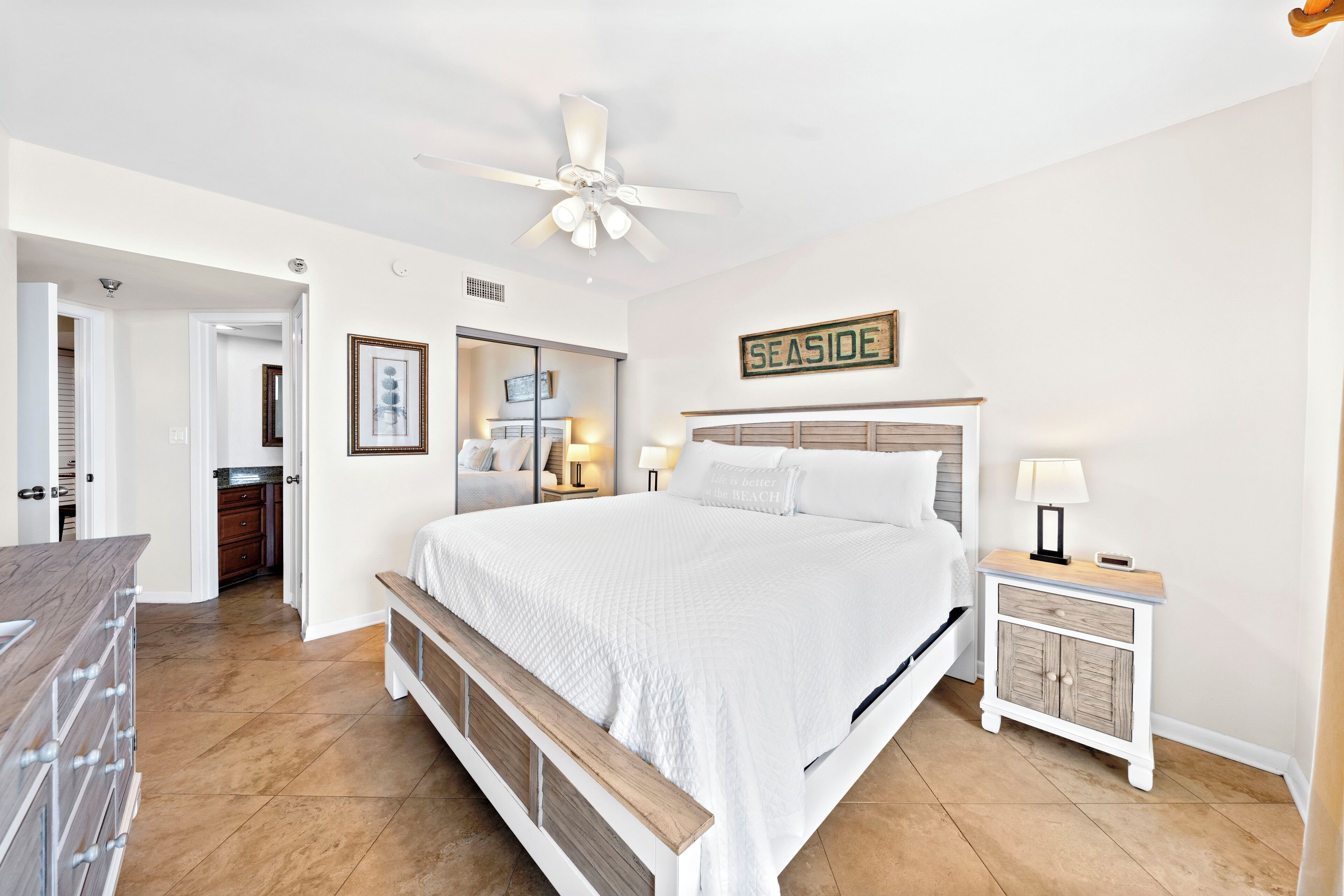 A bedroom with a large bed, a nightstand, a ceiling fan, and a sign that reads 'SEASIDE'.