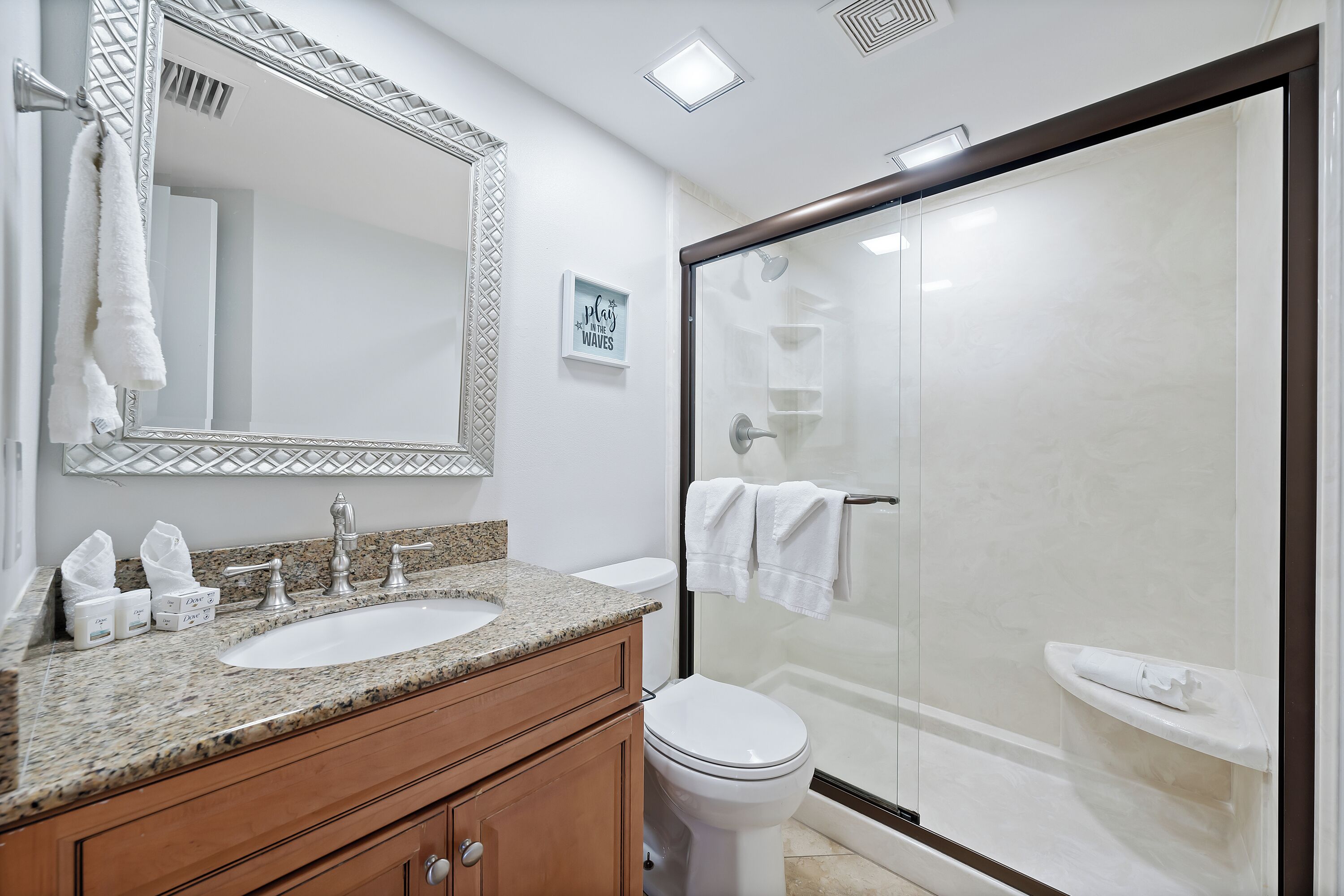 A modern bathroom with a granite countertop, a large mirror, a white toilet, and a glass-enclosed shower.