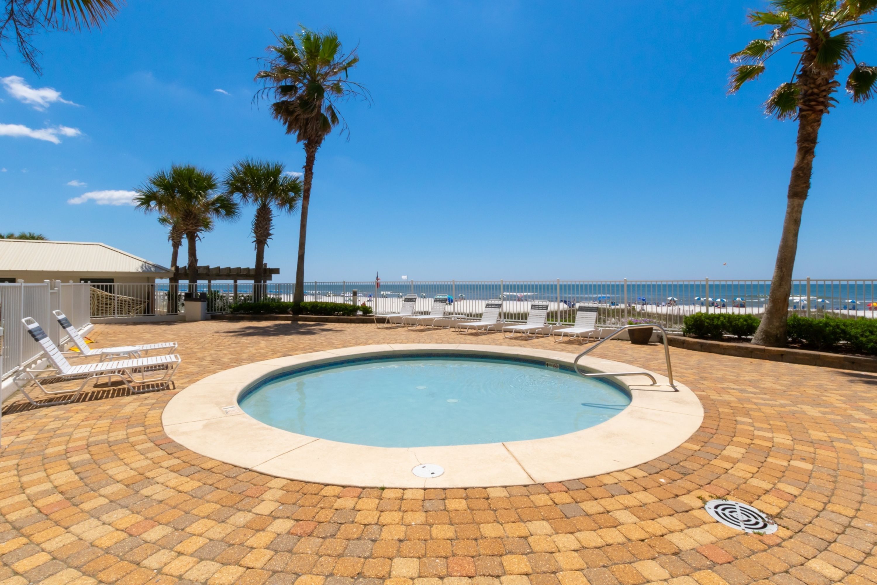 A circular pool with a tiled deck, surrounded by lounge chairs and palm trees, overlooking a beach and ocean.