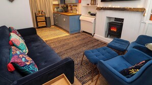 A living room with a sofa, two blue armchairs, a fireplace, a television, and a kitchen area with blue cabinets and a coffee machine. - Seahorse Cottage (St Ives)