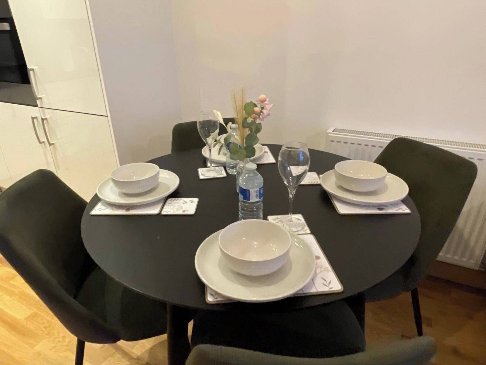 Una mesa redonda de comedor con cuatro platos blancos, dos botellas de agua y un florero con flores.