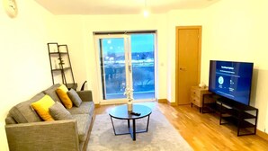 A living room with a grey sofa, a coffee table, a TV on a stand, a large window with a view, and a wooden floor. - Dartford Apartment - Sleeps 3 - Central Stay (Dartford)