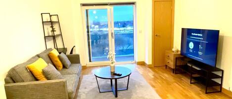 A living room with a grey sofa, a coffee table, a TV on a stand, a large window with a view, and a wooden floor.