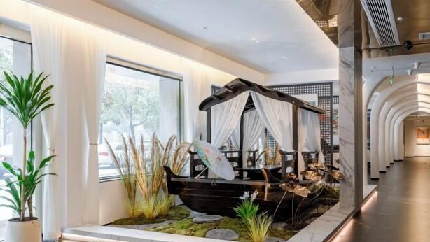 A modern hotel lobby with a large window, a decorative boat centerpiece, and potted plants. - Hehuaju Hotel (Chuzhou Suning Plaza) (Chuzhou)