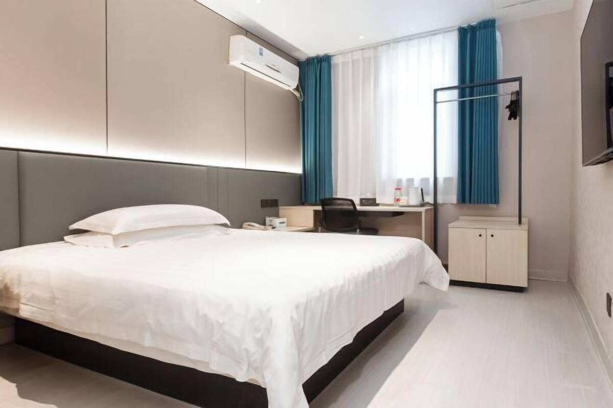 A modern hotel room with a large bed, a desk, a chair, and a wardrobe.