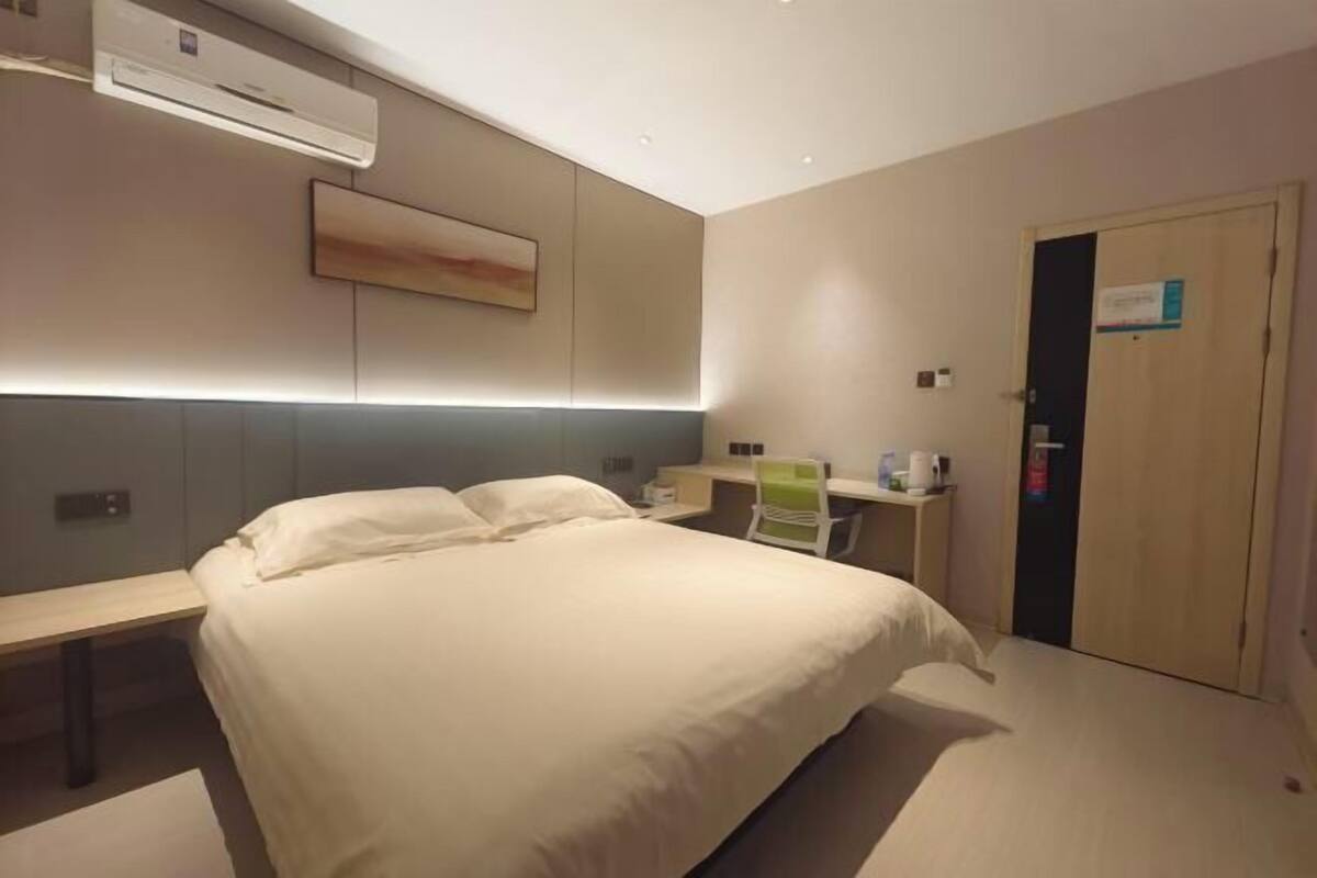 A hotel room with a bed, bedside table, desk, chair, and an air conditioning unit.