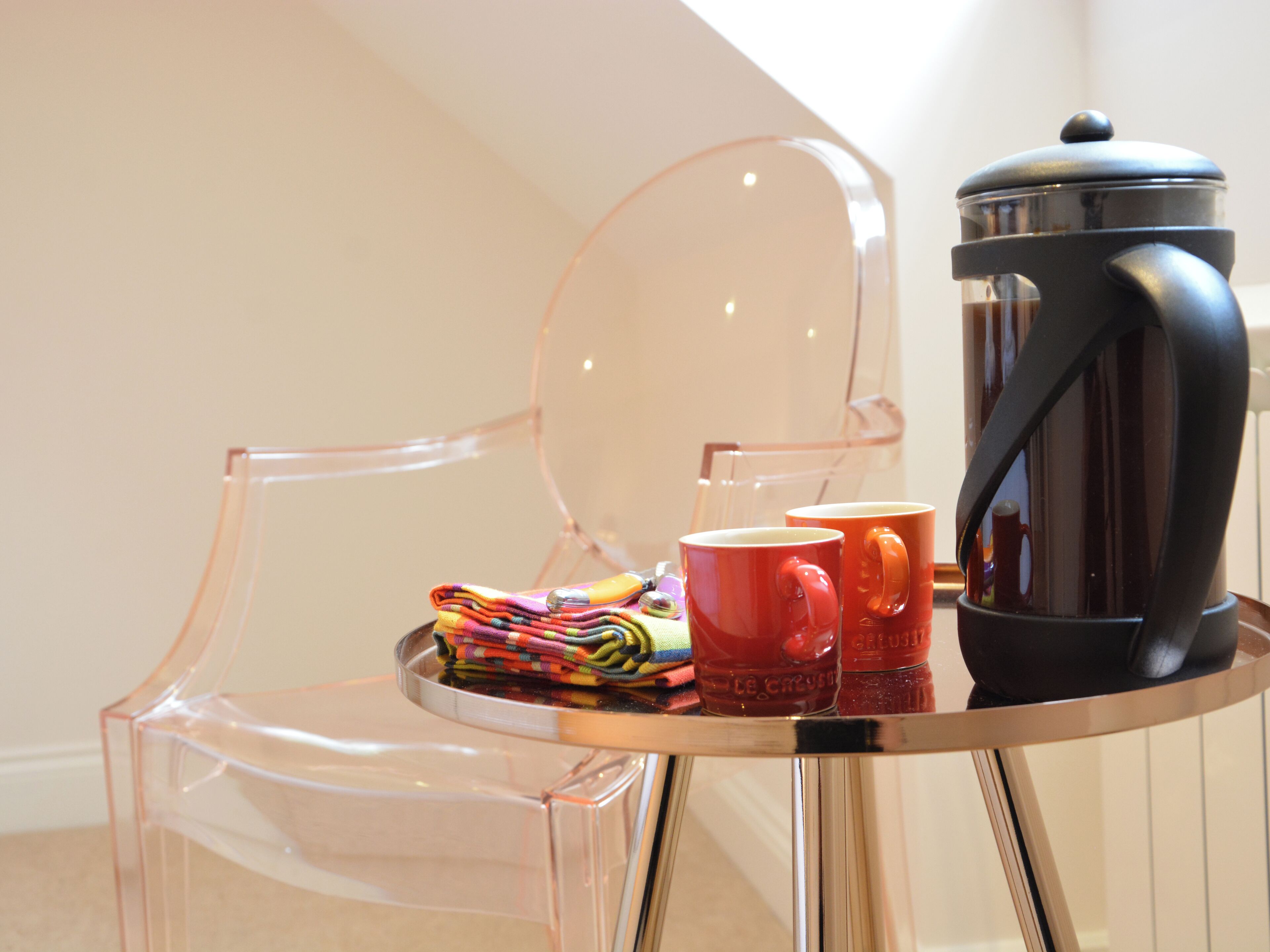 A clear glass chair with a metal table beside it, holding a coffee pot, two red mugs, and a colorful cloth.
