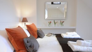 Una cama bien hecha con almohadas blancas y naranjas, una manta de color negro y toallas blancas dobladas cuidadosamente.