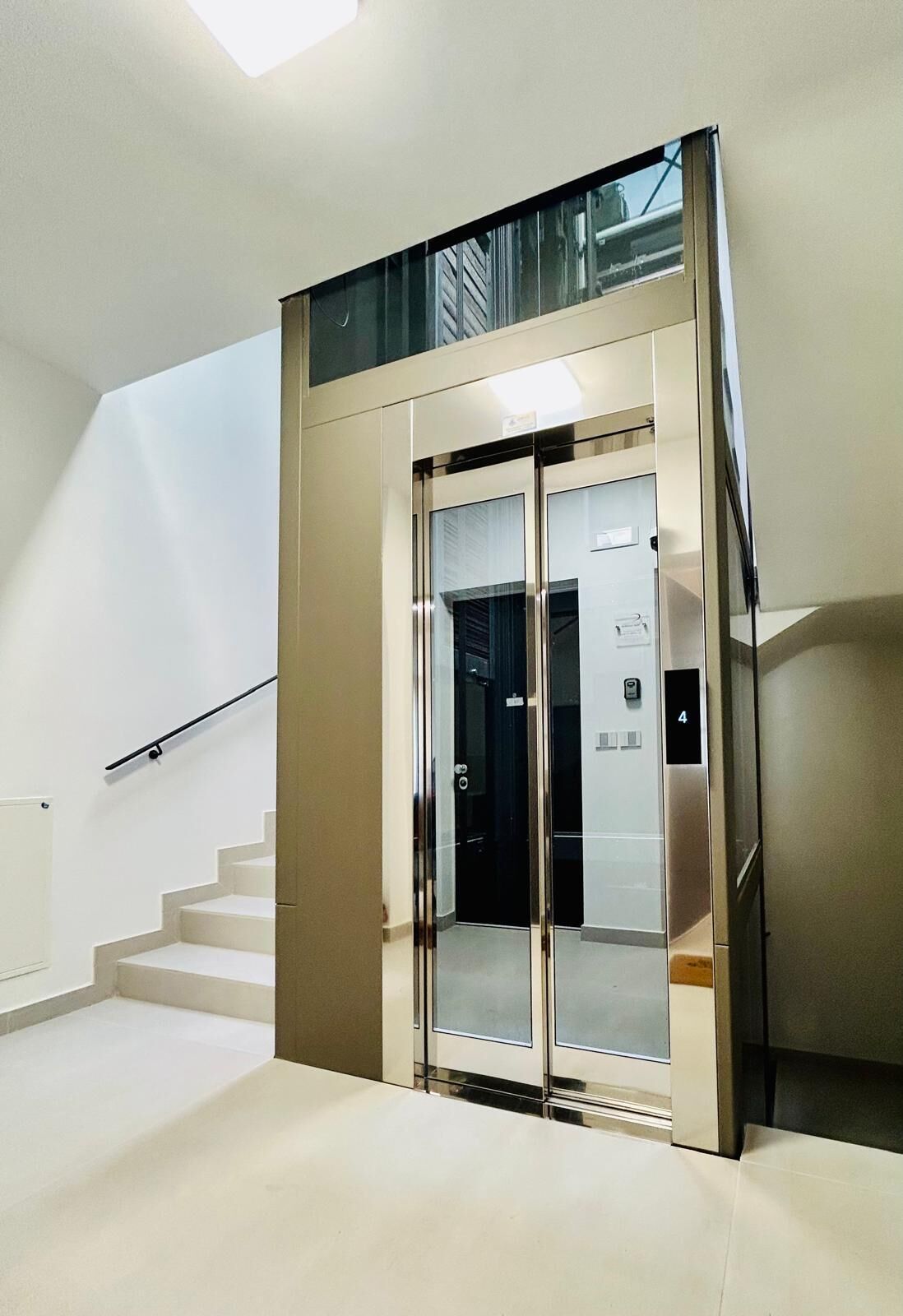 A modern elevator with a glass door, a staircase with a black handrail, and a number 4 sign on the wall.