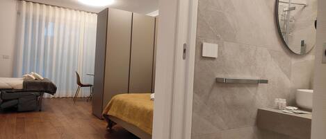 A bedroom with a bed, a chair, a massage table, and a bathroom with a toilet, sink, and mirror.