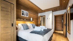 Un dormitorio moderno con techo de madera, una cama grande con ropa blanca y una manta gris. Hay un cuadro en la pared y una mesita de noche con una lámpara.