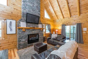 A cozy cabin interior with a stone fireplace, wooden walls, a leather sofa, and a television mounted above the fireplace. - Hocking Creek Reserve | Fenced Yard, Pet Friendly (Logan)