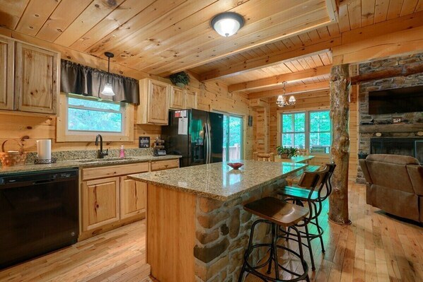 A wooden cabin interior with a kitchen featuring granite countertops, a central island with bar stools, a stone fireplace, and a large window providing natural light. - Buckeye Trail | Hocking Hills, 5BR, Hot Tub, Pets (Rockbridge)