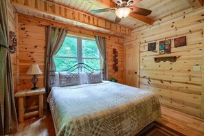 A wooden cabin bedroom with a bed, curtains, a window, a ceiling fan, and wall decorations.