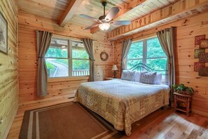 A wooden cabin bedroom with a bed, curtains, a ceiling fan, and a window with a view of greenery. - Buckeye Trail | Hocking Hills, 5BR, Hot Tub, Pets (Rockbridge)