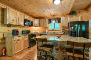 A wooden cabin kitchen with granite countertops, a central island, bar stools, a refrigerator, and a microwave. - Buckeye Trail | Hocking Hills, 5BR, Hot Tub, Pets (Rockbridge)